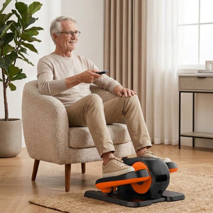 ⁠Senior man sitting in an armchair using a compact under-desk elliptical exerciser while holding a remote control.⁠