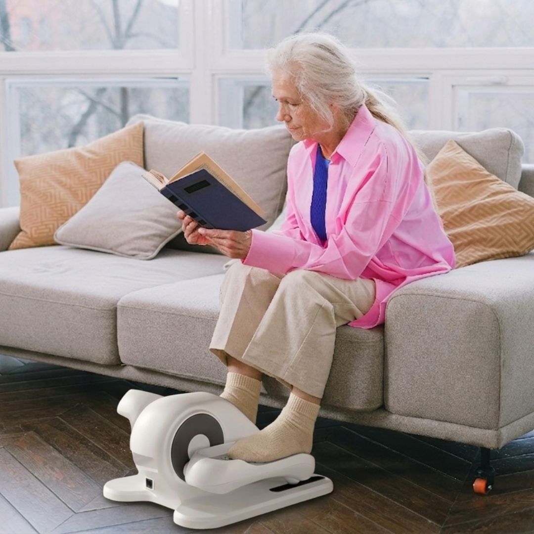 Senior woman in a pink shirt reading a book while exercising with a white under-desk elliptical machine on a living room sofa.