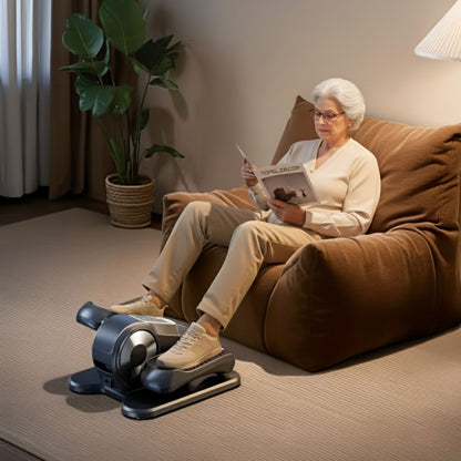 Woman using a mini elliptical black reading a magazine on a brown couch.