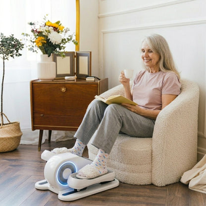 Senior woman using a white under-desk elliptical machine while reading a book and drinking tea in a cozy living room.